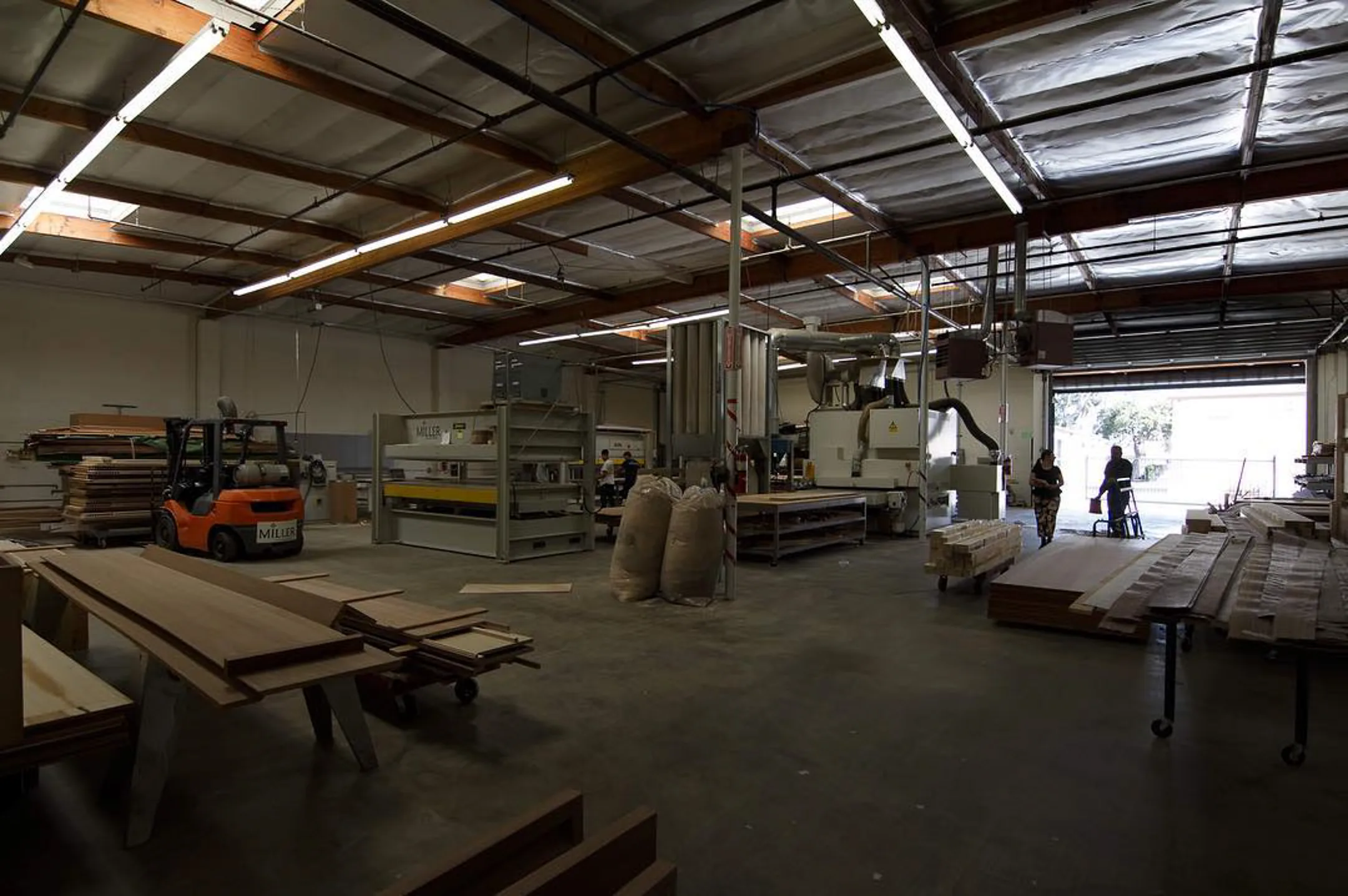 The veneer shop bay from one end: the press and the sander down the middle of the floor, the roller door open to daylight at the far end.