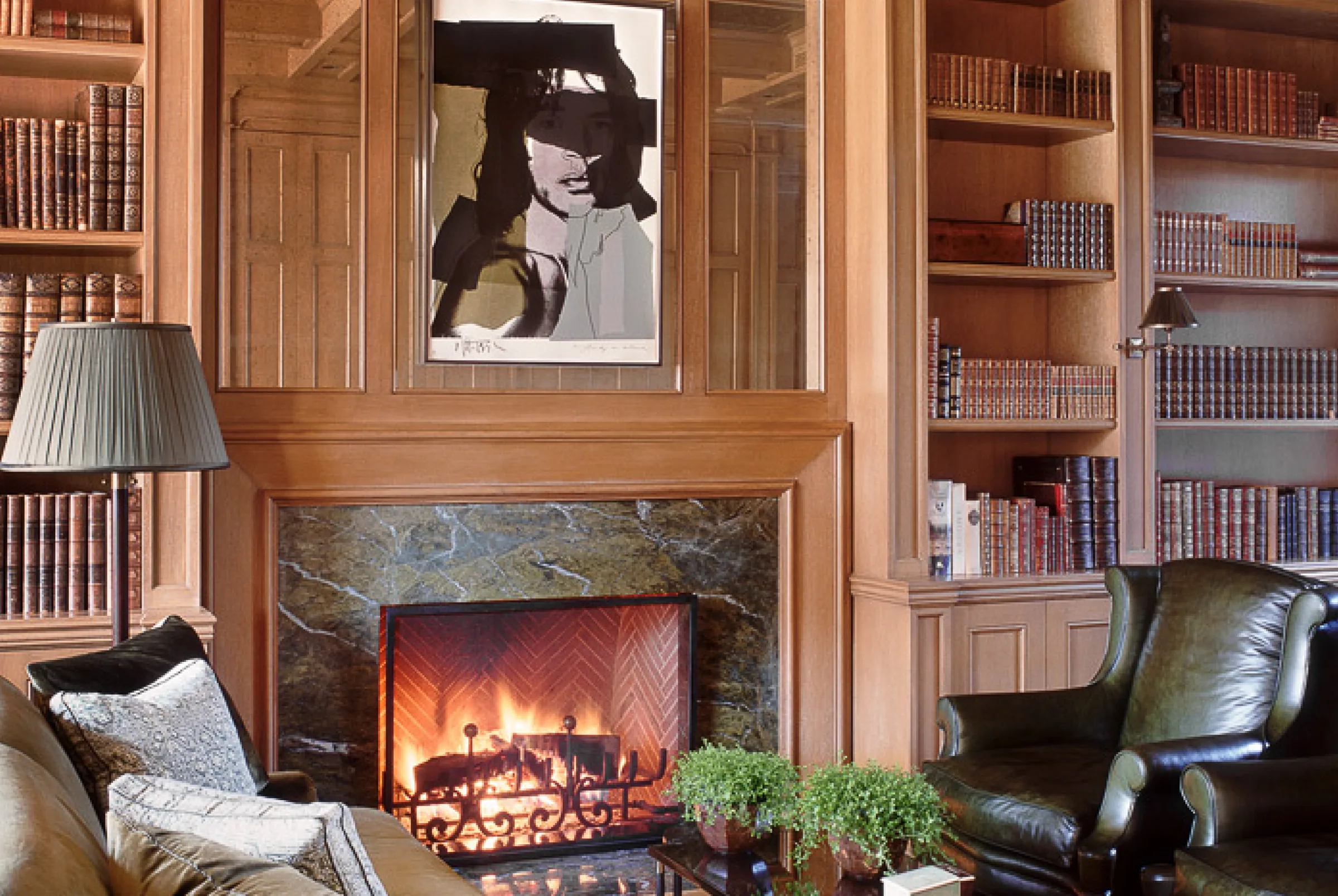 A panelled library in walnut, bookshelves either side of a stone fireplace, leather chairs drawn up to it.