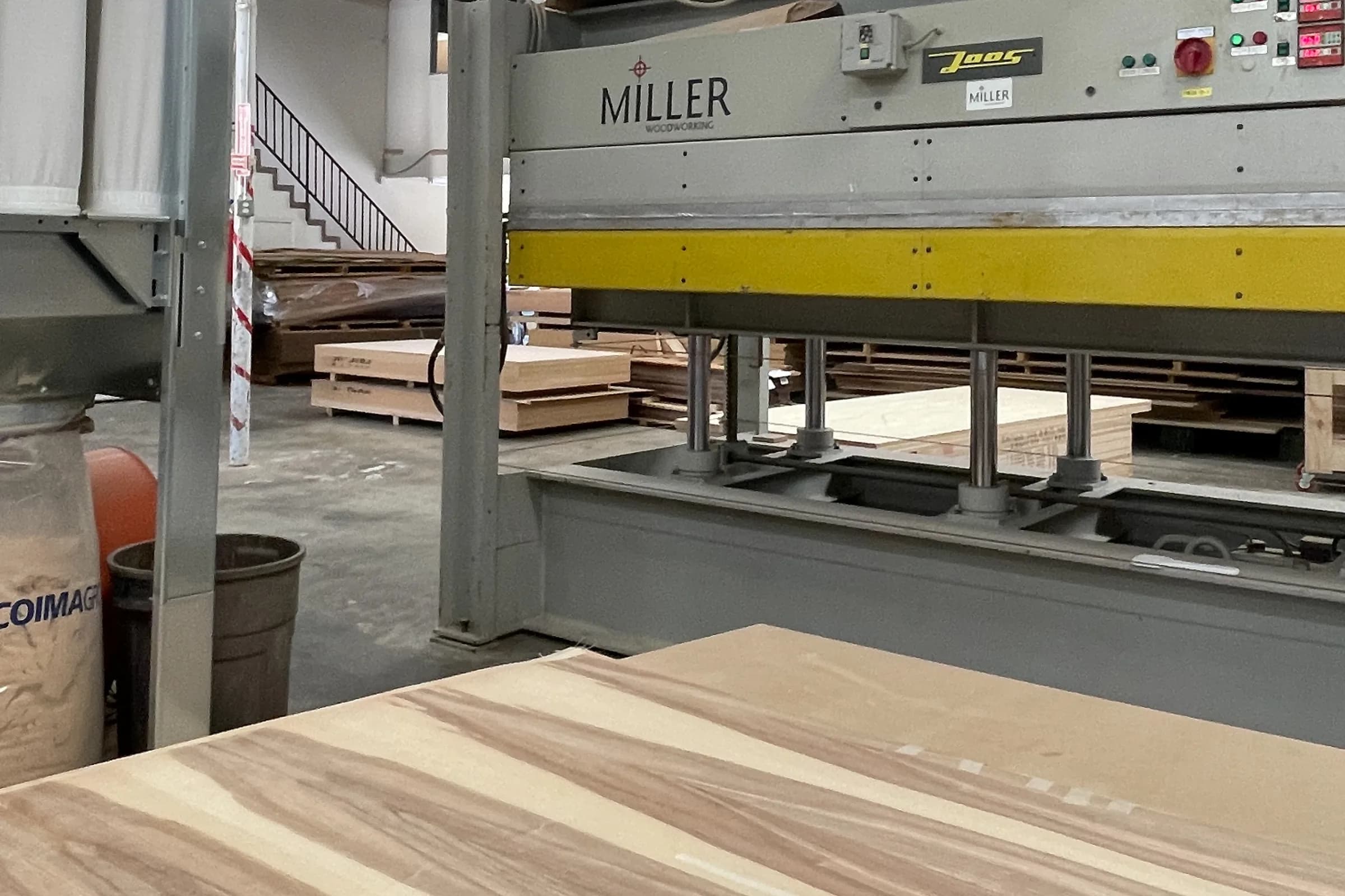 A pressed panel of figured veneer on the bench in front of the hot press, with rack stock and the shop floor behind.