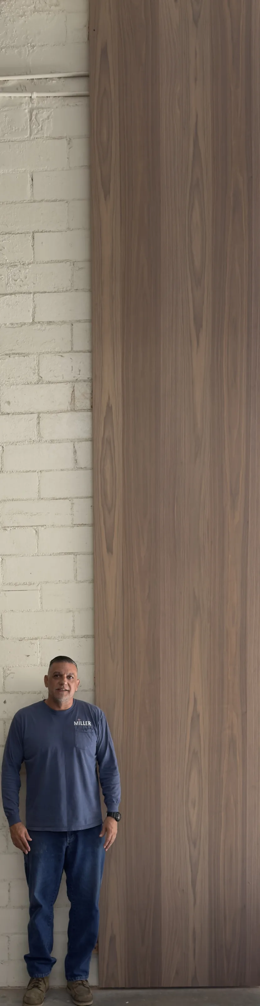 An eighteen-foot walnut panel standing on end against the shop's white brick wall, a man beside it for scale.