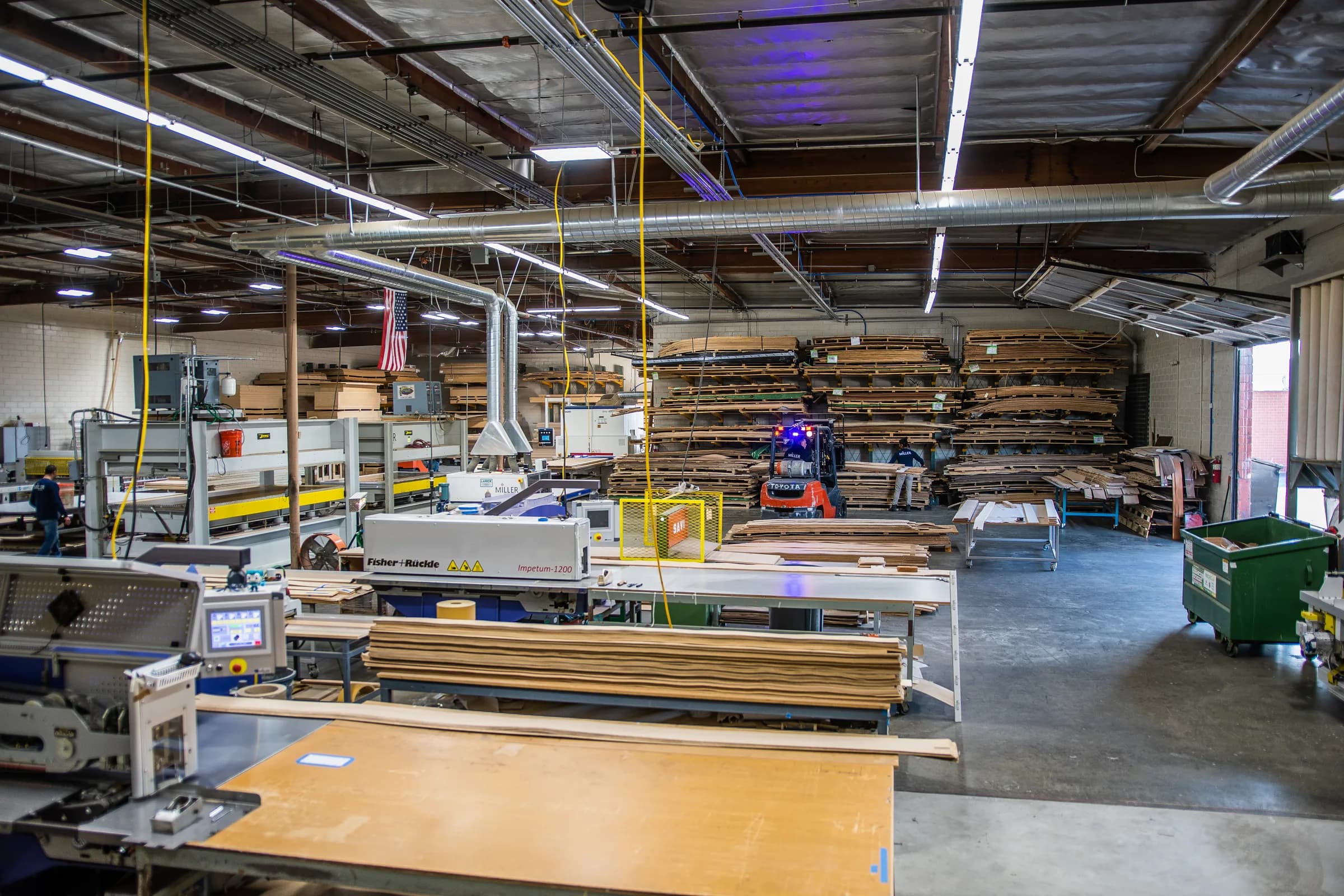 The veneer shop floor from the bench: the press at center, racks of veneer beyond, faces laid out on the tables.